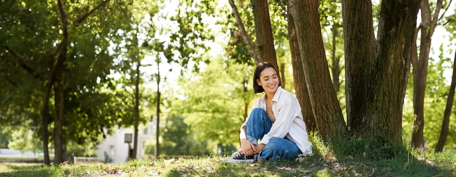 Portrait of beautiful asian woman resting near tree, relaxing in park, smiling and looking happy. Copy space