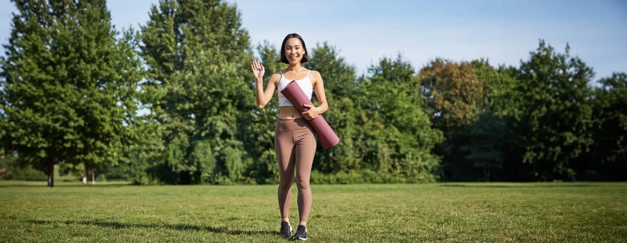 Stylish sportswoman with rubber mat for yoga, says hello, waves at you, ready for workout training outdoors in park on lawn.