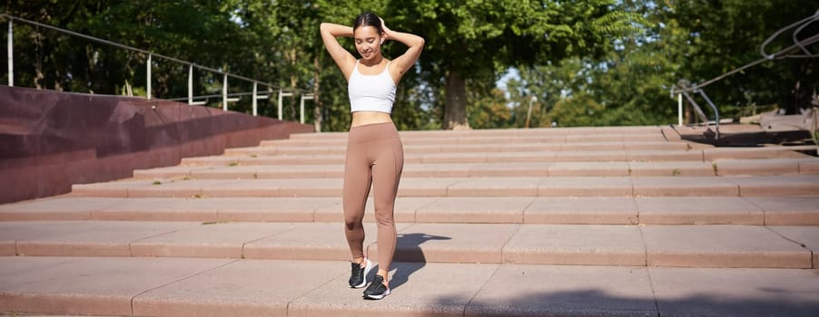 Sportswoman jogging outdoors. Smiling asian fitness girl in wireless earphones, listening music and running on road, workout.