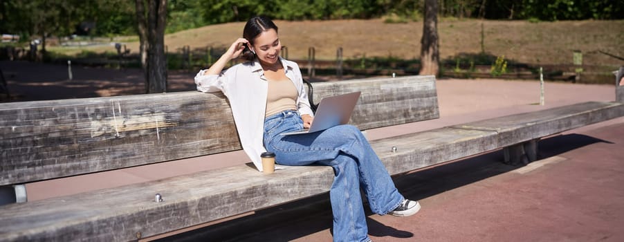 Smiling korean girl sitting in park with laptop, drinking takeaway coffee, enjoying sunny day, working or studying outdoors.