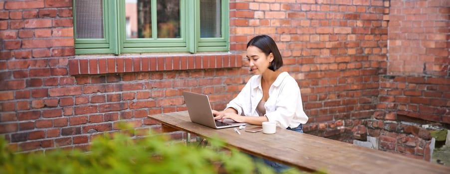 Stylish young urban girl in outdoor cafe, sitting on bench with laptop, smiling, browing on computer.