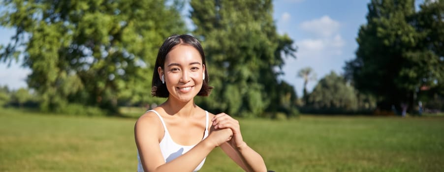 Vertical shot of young fit woman does squats in park, using stretching band on legs, smiling pleased while workout.