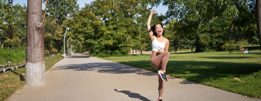 Happy asian girl celebrating victory, saying yes, finish running, workout in park with excitement and joy.