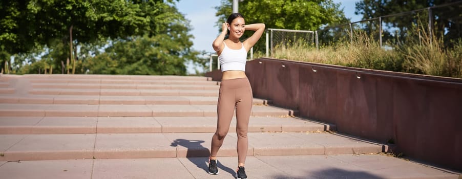 Sportswoman jogging outdoors. Smiling asian fitness girl in wireless earphones, listening music and running on road, workout.