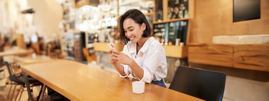 Vertical portrait of stylish asian woman sitting in cafe, drinking coffee and using smartphone. Lifestyle and people concept