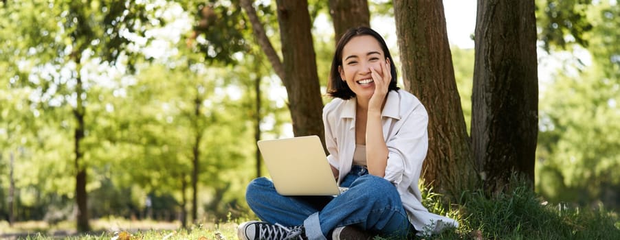 Portrait of young asian woman sitting in park near tree, working on laptop, using computer outdoors.