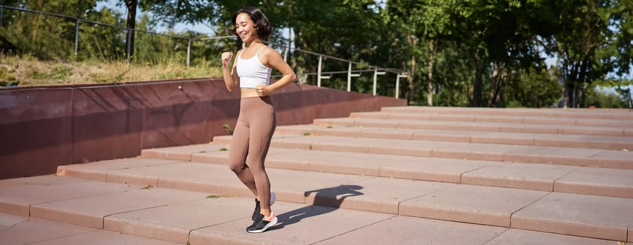 Portrait of young sportswoman running on street. Fitness girl jogging in park, workout outdoors, listening music in wireless earphones.