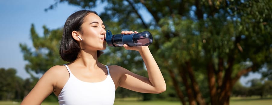 Smiling asian fitness girl drinks water, workout in park, stay hydrated during jogging training session.