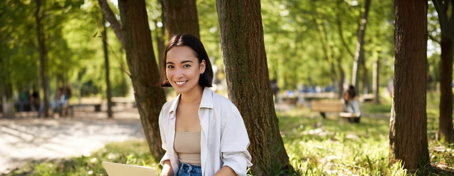 Beautiful asian girl sitting in park with laptop, working on remote, typing on keyboard, smiling at camera, resting beside tree.