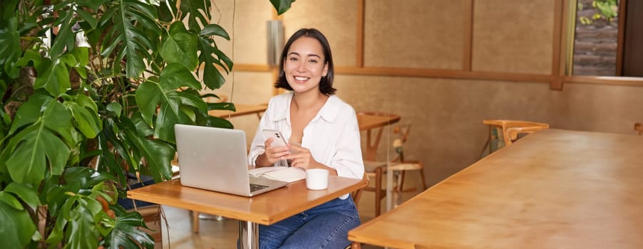 Vertical shot of young asian girl sits in cafe with laptop and smartphone, relaxing and surfing the net, working remotely.