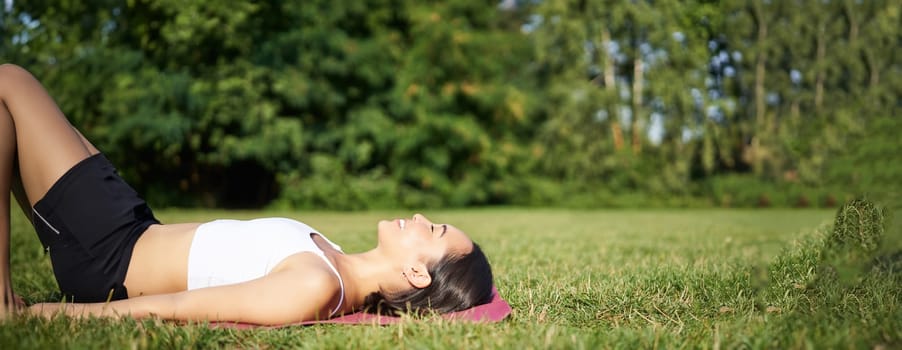 Young fitness girl lying on sport mat on lawn, breathing and meditating in park in sportswear.