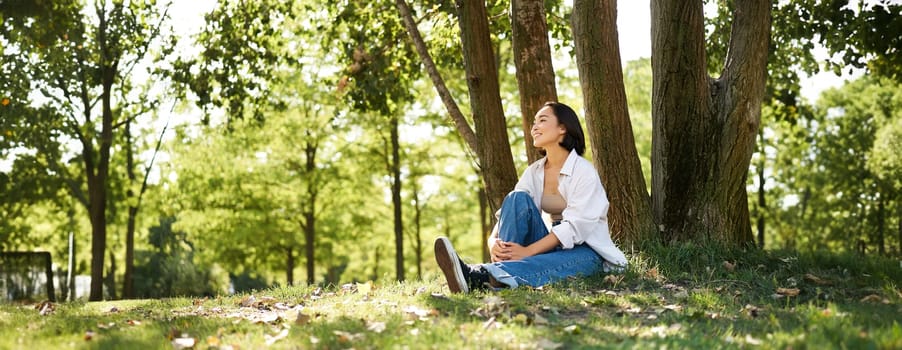 Portrait of asian girl relaxing, leaning on tree and resting in park under shade, smiling and enjoying the walk outdoors.