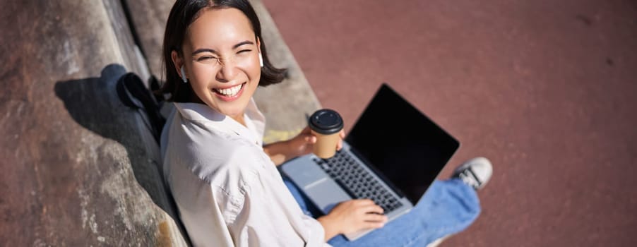 Beautiful young asian female student, sitting with laptop on bench in park, drinking takeaway coffee and listening music, working remotely, studying outdoors.