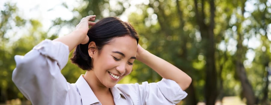 Beautiful young woman tying her hair while walking in park, smiling romantic, enjoying warm day.