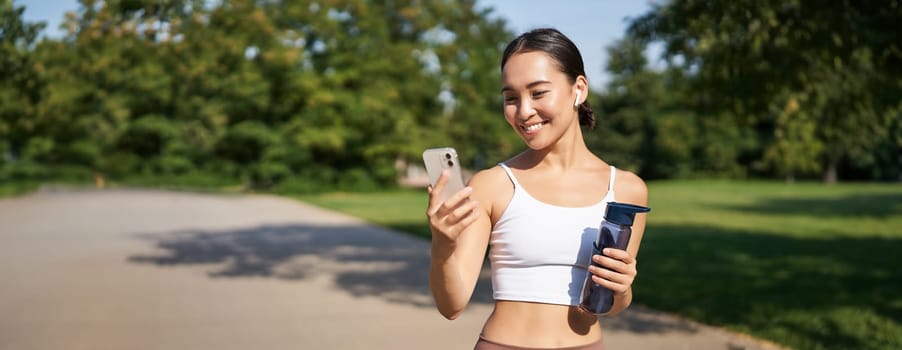 Fitness woman with water bottle and smartphone, jogging in park and smiling, looking at her mobile phone app, checking sport application.