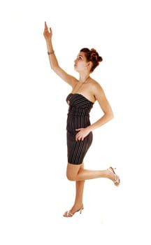 A pretty and young woman in a black dress with stripes on standing from the side for white background with one hand the ear,.