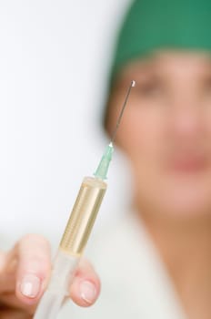 Close-up of a plastic syringe filled with serum with sharp drops at the tip of the needle in the hand of a female doctor dressed in a green surgical cap in blurred background