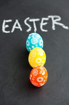 Closeup of three colorful Easter egg shaped candles and text written in chalk on dark background