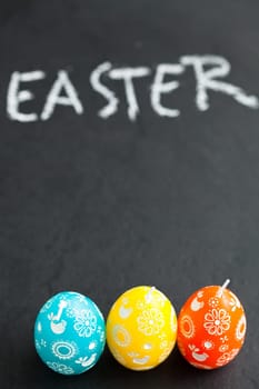 Closeup of three colorful Easter egg shaped candles and text written in chalk on dark background