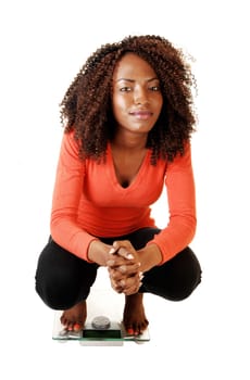 A lovely black teen girl crouching on a scale, looking into the camera, for white background in an orange sweater and black tights.