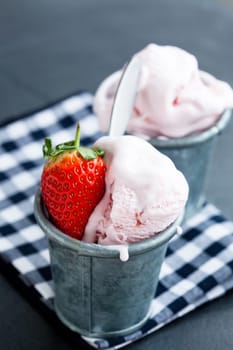 Closeup of strawberry ice cream and strawberry in two metal cups on checkered serviette over dark background