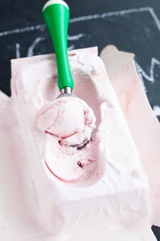 Closeup of tub of strawberry ice cream and scoop on dark background