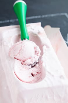 Closeup of tub of strawberry ice cream and scoop on dark background