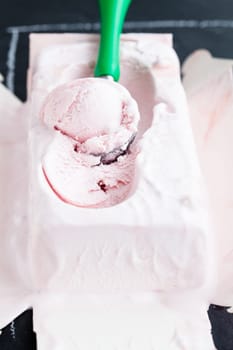 Closeup of tub of strawberry ice cream and scoop on dark background