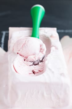 Closeup of tub of strawberry ice cream and scoop on dark background