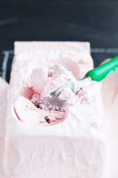 Closeup of tub of strawberry ice cream and scoop on dark background