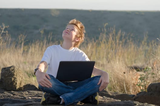 Handsome kid with laptop sits in nature and look at the sky