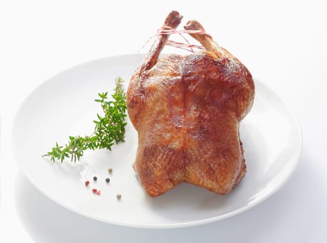 Whole crisp golden roast duck served on a plain white plate with a sprig of fresh rosemary over a white background