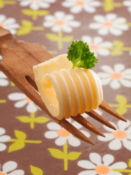 Decorative curl of rolled butter or margarine garnished with fresh mint being offered on a wooden salad fork with a floral tablecloth background