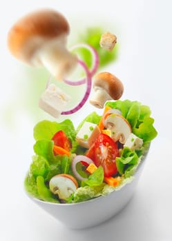 High angle view of a bowl of healthy fresh mixed salad with lettuce, tomato, cheese, onion and sliced mushrooms over a white background with blurred ingredients