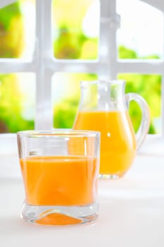 Healthy freshly squeezed orange juice in a glass tumbler with a glass jug full of juice standing in front of a fresh summery window