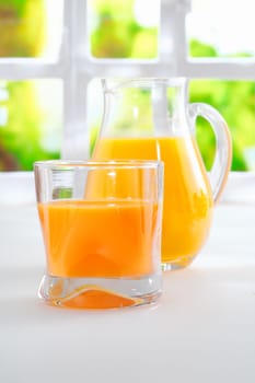 Tumbler of fresh healthy orange juice standing on a kitchen table alongside a jug full of juice with a sunny window and greenery behind