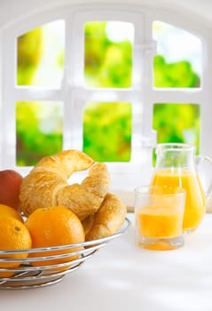 Wire basket filled with fresh whole oranges and croissants for a summer breakfast standing in front of a sunny window with greenery