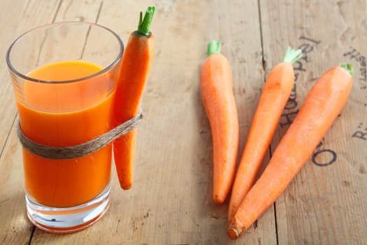 A tall glass of healthy freshly blended carrot juice on a wooden table with a whole carrot attached and a trio of raw carrots lying alongside