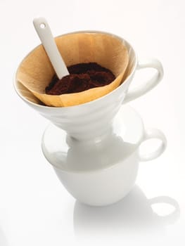Portable plain white china filter funnel with filter paper filled with freshly ground coffee beans and a measuring spoon
