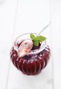 Elegant fluted glass jar of berry jam with a used silver teaspoon coated in jam resting on top of the rim with three fresh mint leaves