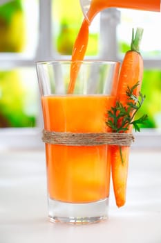 Pouring fresh carrot juice from a jug into a glass with a raw whole carrot strapped to the side with string