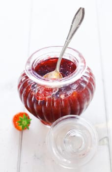Delicious homemade strawberry preserve in an elegant fluted glass jar with a silver serving spoon on a white background