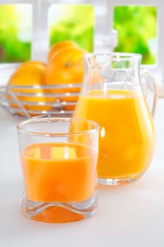 Freshly squeezed orange juice for breakfast standing on a white tabletop with a full glass tumbler and jug and a bowl of fresh oranges in the background against a sunny window with greenery