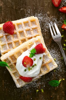 Delicious waffle topped with whipped cream and ripe red fresh strawberries sprinkled with sugar and served on a rustic wooden surface