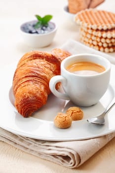 Freshly baked crisp golden croissant served with a cup of rich espresso and two small macaroon biscuits in a restaurant