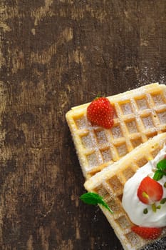 Overhead view of golden waffles with cream and strawberries on a rough textured wooden table with copyspace