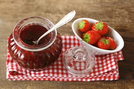 High angle view of a rustic still life with fresh strawberries and strawberry jam in a pretty glass jar arranged on a cheerful red and white checked cloth on an old wooden tabletop