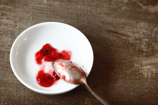 Silver teaspoon resting on a small white dish with samples of fresh fruity strawberry jam taken while preparing a batch in the kitchen