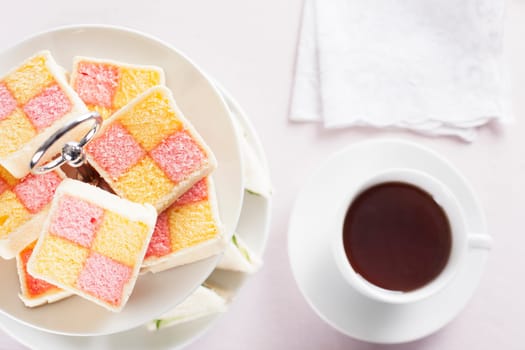 Battenberg cake on a two tiered serving plate and a cup of tea on the table