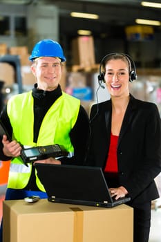 Teamwork - warehouse clerk or warehouse worker and customer service representative using laptop and headset in a warehouse of a freight forwarding company,she points at the camera